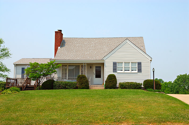A single-story house with light gray siding, dark shutters, and a brick chimney, sits on a neatly trimmed lawn with bushes and a wooden deck on the left side.